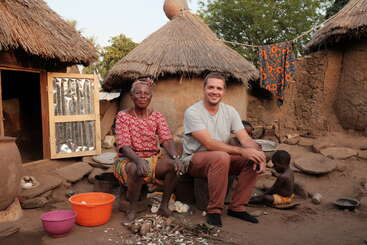 An elderly woman and a man sit smiling outside traditional thatched huts, with children nearby. Bowls, utensils, and vibrant clothing create a warm, rustic village scene.