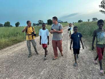 Eine Gruppe von Menschen, darunter Erwachsene und Kinder, spaziert tagsüber gemeinsam auf einem Schotterweg durch grasbewachsene Felder, unterhält sich dabei und erkundet die Umgebung.