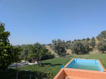 The image shows a peaceful countryside garden with green grass, tall trees, a blue sky, and an above-ground swimming pool, surrounded by nature and hills.