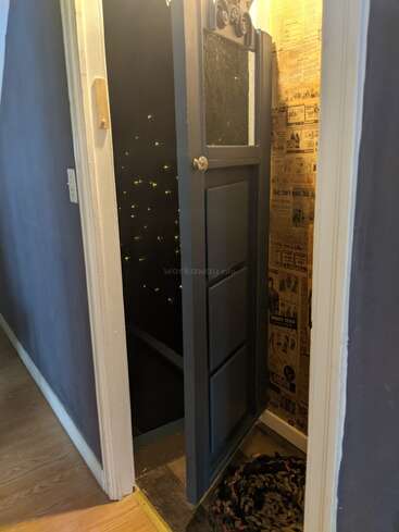 Un escalier étroit et mystérieux se cache derrière une porte bleu foncé, éclairé par de petites guirlandes lumineuses. Les murs sont décorés d'imprimés de journaux vintage, créant une atmosphère intrigante.