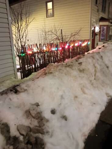 Une grande pile de neige sale se trouve devant une clôture en bois décorée de lumières de Noël colorées, à côté de maisons jaunes, par une froide nuit d'hiver.