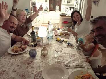 Uma cena alegre de jantar em família com cinco adultos e uma criança, sorrindo e levantando as mãos, sentados em torno de uma mesa festivamente posta em uma cozinha com iluminação aconchegante.