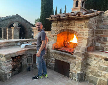 Um cara está sorrindo enquanto grelha carne em um forno rústico de pedra ao ar livre com uma fogueira crepitante, cercado por lenha empilhada e uma bela arquitetura rural.