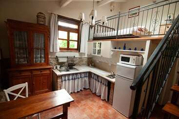 Essa cozinha aconchegante tem móveis rústicos de madeira, uma área com cama alta, armários brancos, piso de azulejo, uma pequena mesa de jantar, decoração vintage e luz natural que entra pelas janelas.