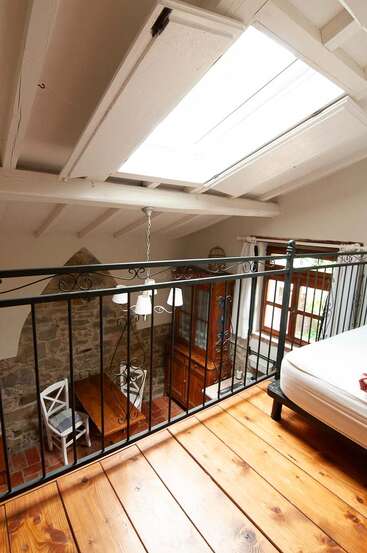 Esse loft aconchegante tem piso de madeira polida, corrimão de ferro forjado, parede de pedra exposta, claraboia, área de jantar rústica com cadeiras brancas e luz natural quente.