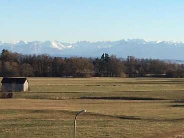 L'image représente un paysage serein composé d'un champ, d'un petit bâtiment, d'arbres et d'une majestueuse chaîne de montagnes sous un ciel bleu clair.