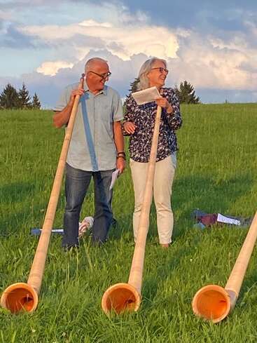 L'image représente un homme et une femme debout dans un champ herbeux, chacun tenant un cor des Alpes, avec trois autres cornes visibles au premier plan.