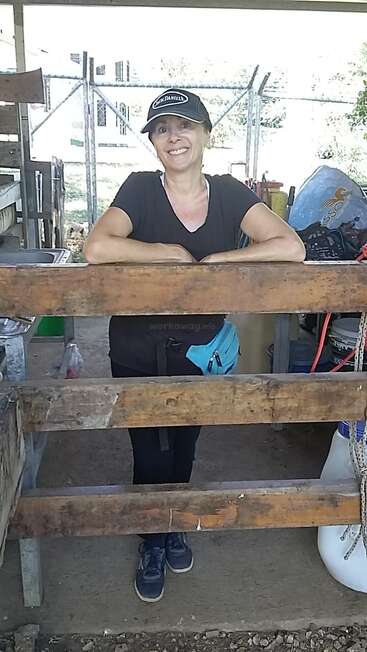 Eine lächelnde Frau in einem schwarzen Shirt, einer Mütze und einer türkisfarbenen Gürteltasche lehnt an einem Holzzaun in einem rustikalen Arbeitsbereich im Freien, hinter ihr stehen verschiedene Geräte.