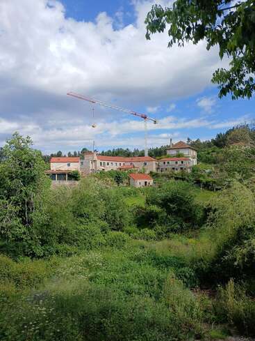 L'image représente un paysage serein avec un grand bâtiment au toit rouge, entouré d'une végétation luxuriante et d'une grue, sur fond de ciel bleu avec des nuages blancs.