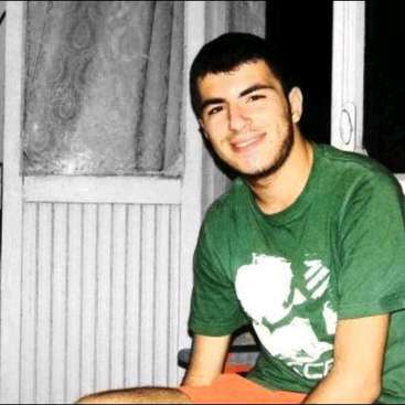 The image depicts a young man with short dark hair, wearing an olive green t-shirt with a white design, sitting in front of a door with a window and curtains.