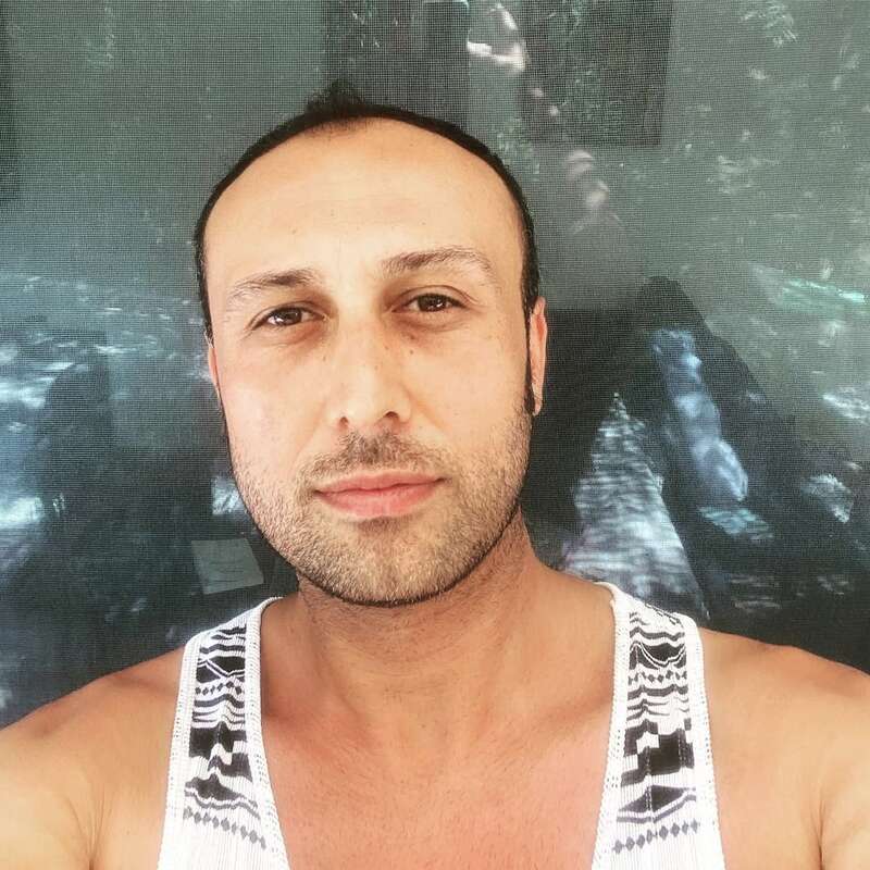 La imagen muestra a un hombre con barba corta y bigote, vestido con una camiseta de tirantes blanca con dibujos negros, sobre un fondo gris con textura.