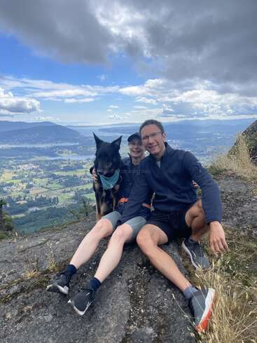 Two people and a black dog sit on a rocky hilltop, smiling, with a breathtaking view of mountains, valleys, and cloudy blue sky behind them.