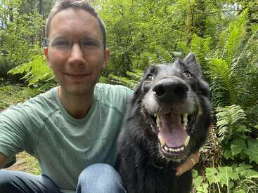 A man wearing glasses and a green shirt smiles beside a happy black dog with its mouth open, both enjoying a peaceful moment in a lush forest.