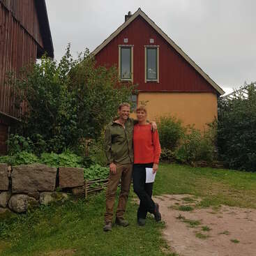 Ein Mann und eine Frau stehen lächelnd zusammen in einem Garten, mit Grünzeug und einem rot-gelben Haus hinter ihnen, unter einem grauen, bewölkten Himmel.