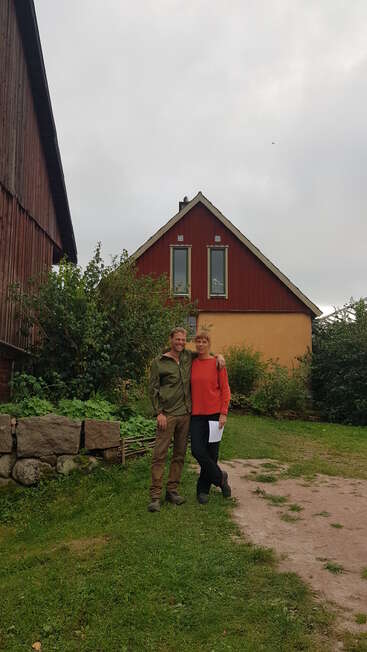 Un hombre y una mujer sonríen juntos en un jardín, con vegetación y una casa roja y amarilla detrás de ellos, bajo un cielo gris y nublado.