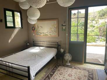 Um quarto aconchegante com uma cama de metal preta, lanternas de papel, tons terrosos, quadros na parede, um pequeno ventilador, uma porta aberta com vista para a vegetação e a luz natural do sol entrando pelo janelo.