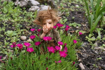 Das Bild zeigt eine kleine, handgefertigte Trollpuppe mit braunen Haaren und Ohren, die inmitten von rosa Blumen steht und ein grünes Hemd und eine braune Hose trägt.
