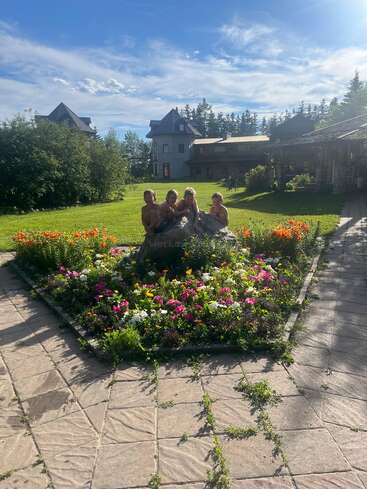 Quatre enfants sont assis sur un rocher au milieu d'un parterre de fleurs coloré, entourés d'un jardin avec des maisons pittoresques et un ciel bleu éclatant.