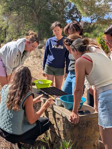 A group of women gathers outside around a stone table, watching a demonstration involving soil, plants, and containers, possibly participating in a gardening or outdoor educational activity.