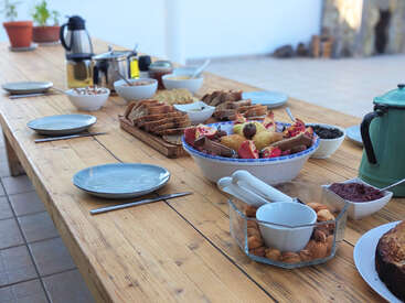 A long wooden table is set outdoors with plates, bread, fruits, coffee, pastries, and spreads, ready for a hearty breakfast or brunch gathering. Inviting atmosphere.