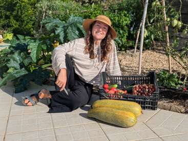 A woman wearing a hat sits happily in a garden with a basket of fresh vegetables and fruits, and two large zucchinis, enjoying the sunshine.