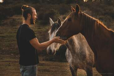 Um homem de barba e coque alimenta ou acaricia gentilmente dois cavalos ao ar livre ao pôr do sol, criando um momento sereno e acolhedor de conexão com a natureza.