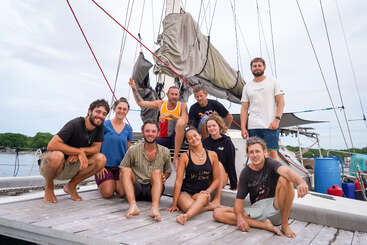 Un grupo de nueve personas, sonrientes y relajadas, posan juntas en la cubierta de un velero. El fondo muestra agua, vegetación y las jarcias del barco.