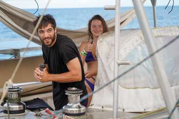Un hombre y una mujer en un velero, sonrientes y relajados. El hombre maneja el equipo y la mujer levanta el pulgar. Al fondo se ve el mar abierto.