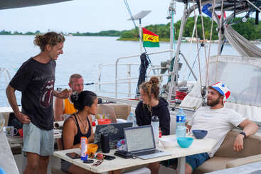 Cinco personas se relajan y conversan en un velero, rodeadas de ordenadores portátiles, comida y bebidas. Se ve una bandera, con agua y vegetación de fondo.