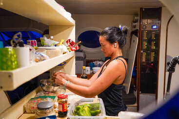 Una mujer prepara comida en una cocina compacta de un barco, rodeada de ingredientes y utensilios, con una camiseta de tirantes negra y auriculares inalámbricos.