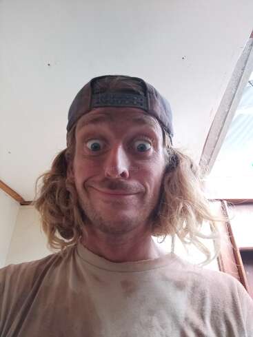 A man with long, messy blond hair, wearing a backward baseball cap and a dirty shirt, smiles widely with wide eyes inside a room with white walls.
