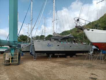 A large catamaran named "Zoe III" is on dry land supported by stands. Other boats and green hills are visible in the background. Sky is partially cloudy.
