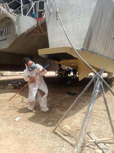 A person wearing protective clothing and a mask is painting the bottom of a large boat on dry land, likely performing maintenance or repairs in a shipyard.