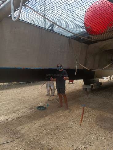 A person wearing a mask stands beneath a large catamaran, painting its hull. Another person in white works nearby. The ground is dusty, tools scattered around.