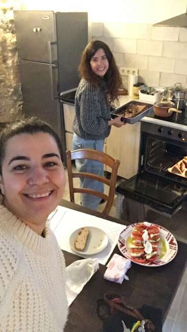 Duas mulheres estão cozinhando juntas, felizes, numa cozinha aconchegante. Uma segura uma bandeja, a outra sorri para a câmera. Comida e pão estão sobre a mesa.