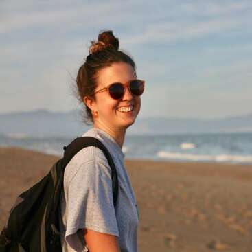 The image depicts a woman standing on a beach, wearing a grey t-shirt, sunglasses, and a backpack, with the ocean and sky visible in the background.