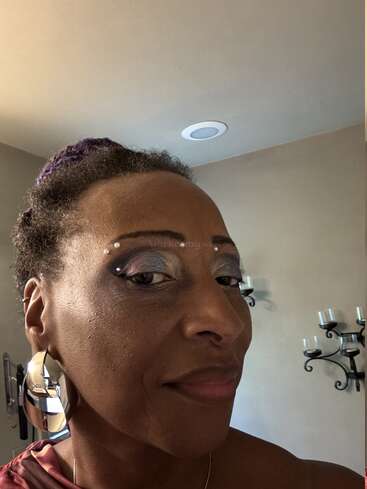 Una persona posa para un primer plano en una habitación bien iluminada, con pendientes metálicos, maquillaje de ojos morado, pedrería y una expresión de confianza. Detrás se ven portavelas.