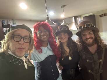 Cuatro personas con disfraces creativos, posiblemente inspirados en el steampunk o la fantasía, sonríen juntas en una cocina cálidamente iluminada. Dos llevan gafas y sombreros, uno una peluca roja.