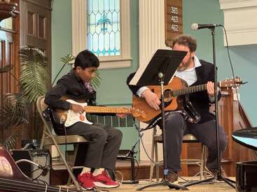 Un niño y un hombre se sientan en sillas y tocan juntos la guitarra en un entorno similar al de una iglesia, con partituras, un micrófono y una vidriera a sus espaldas.