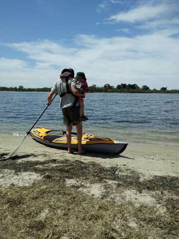 Un homme se tient sur une plage de sable, tenant un enfant et une pagaie, avec un kayak jaune sur le rivage, sur fond d'une étendue d'eau sereine et d'un ciel bleu parsemé de nuages.