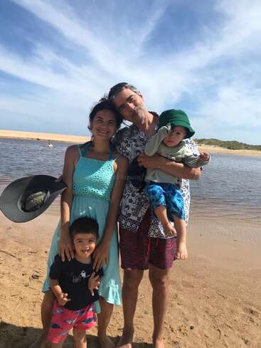 L'image représente une famille de quatre personnes posant sur une plage de sable, avec en toile de fond une étendue d'eau sereine et un ciel bleu parsemé de nuages.
