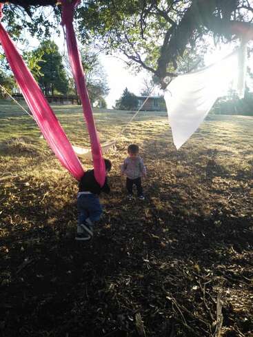 L'image représente deux jeunes enfants jouant dans un champ herbeux et arboré, entourés d'un hamac et d'un drap blanc suspendu à une branche d'arbre.