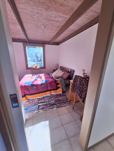 Cette chambre chaleureuse est dotée d'un plafond en bois, d'un couvre-lit coloré, d'un tapis à motifs, d'une fenêtre baignée de lumière, d'une petite lampe et d'une chaise recouverte d'un tissu drapé.