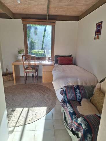 Une pièce chaleureuse et baignée de soleil, avec un lit, un bureau et une chaise. Un grand tapis rond, un petit canapé, la vue depuis la fenêtre et une décoration minimaliste et naturelle créent une atmosphère chaleureuse.
