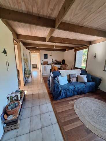 Ce salon chaleureux se distingue par ses poutres apparentes, un canapé bleu orné de coussins, un tapis rond, une décoration éclectique, un sol carrelé et une lumière naturelle abondante provenant de grandes fenêtres.