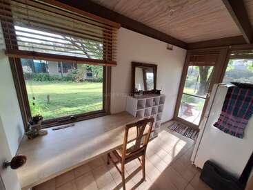 Cette pièce chaleureuse et baignée de soleil comprend un bureau en bois face à une grande fenêtre donnant sur le jardin, une chaise, des étagères, un miroir, un réfrigérateur et une décoration naturelle.