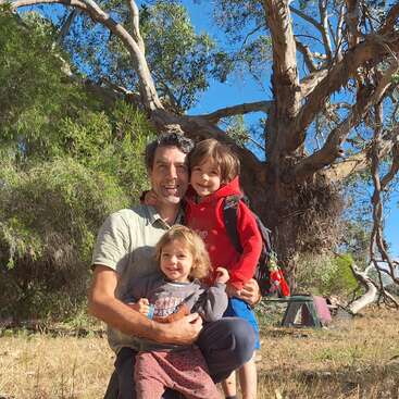 Un homme souriant est agenouillé à l'extérieur, deux jeunes enfants dans les bras. Ils sont entourés d'arbres et d'herbe, profitant d'une journée ensoleillée, peut-être en camping.
