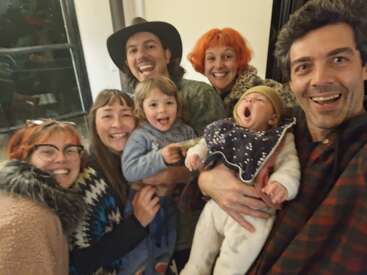 Un groupe de sept personnes, dont deux enfants, sourit joyeusement pour un selfie pris à l'intérieur. Le bébé bâille, capturant un moment spontané, heureux, de convivialité et de chaleur.
