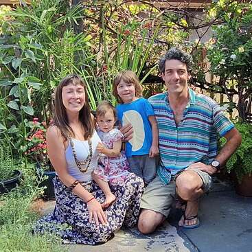 Une famille heureuse de quatre personnes pose en plein air, entourée de plantes et de fleurs luxuriantes. Les deux parents sont agenouillés à côté de leurs enfants souriants, profitant ensemble de la nature.