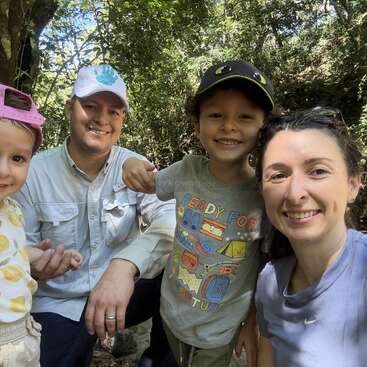 A smiling family of four poses outdoors in a wooded area. The parents and two children wear hats and casual clothes, enjoying a sunny nature adventure together.